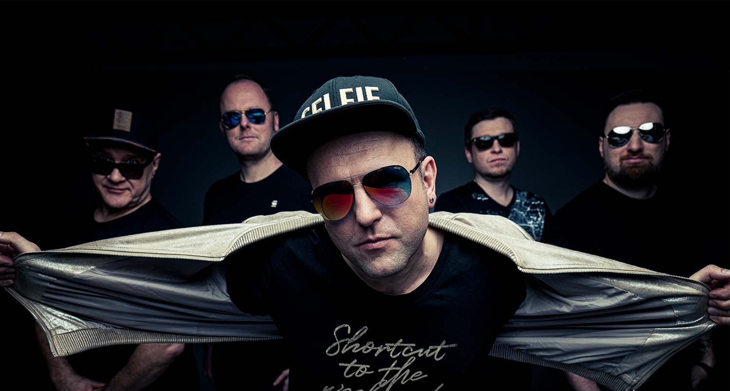Dunkles Pressefoto der Band. Sänger im Vordergrund mit Sonnebrille und Cappy, Band im Hintergrund.